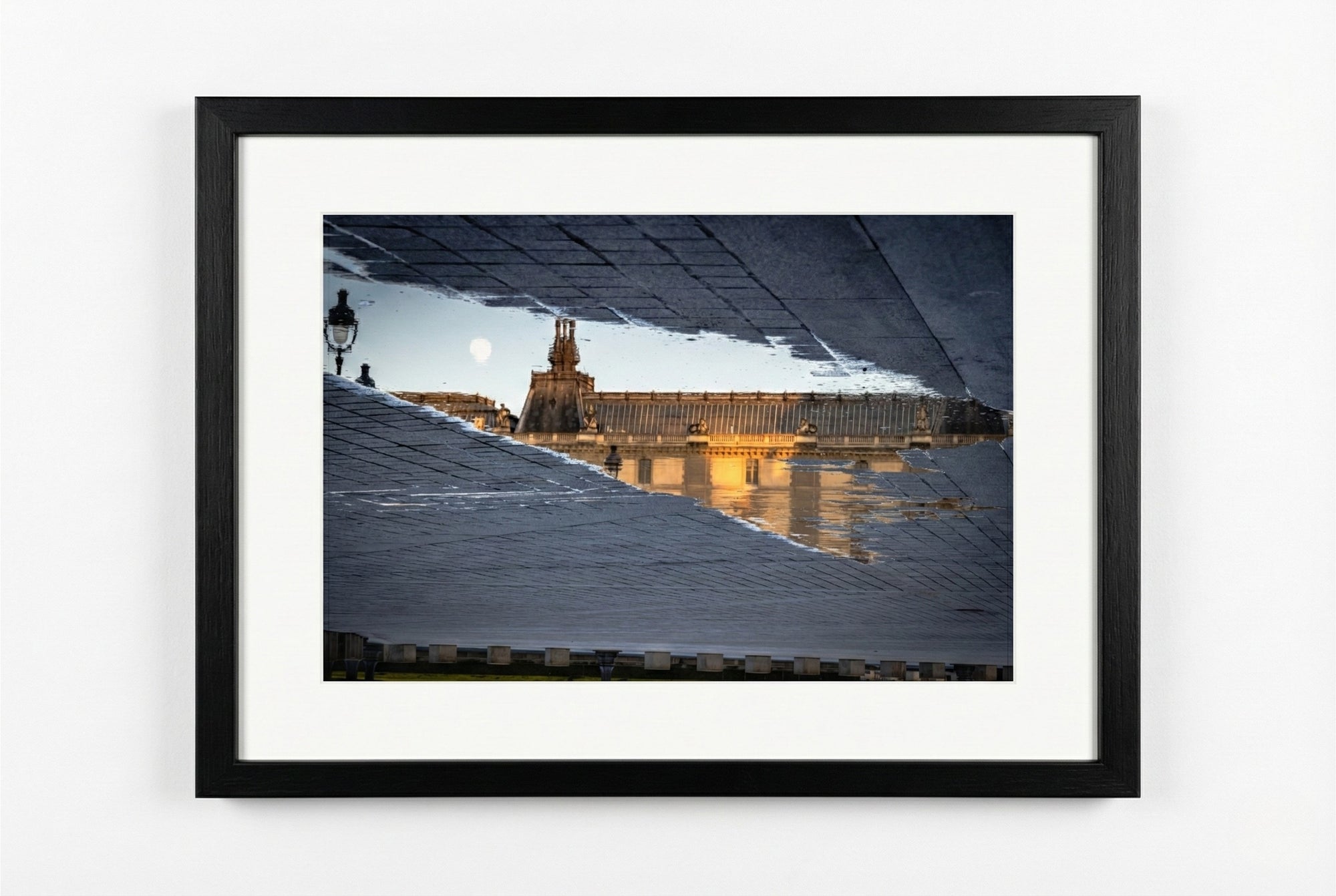 Framed photograph of a cityscape with a large building and street lamps on a white wall.