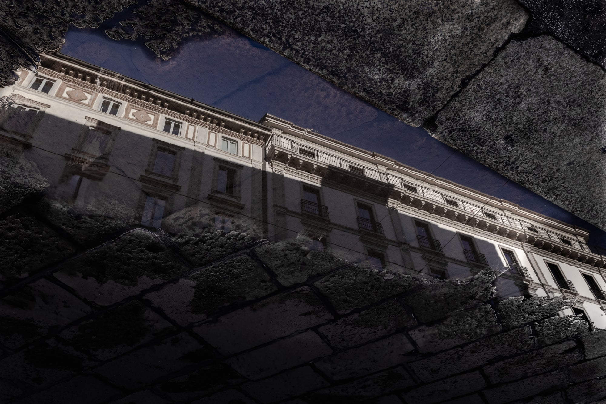 Reflective surface from a puddle on a cobblestone street showing a building's reflection with a dark, textured background